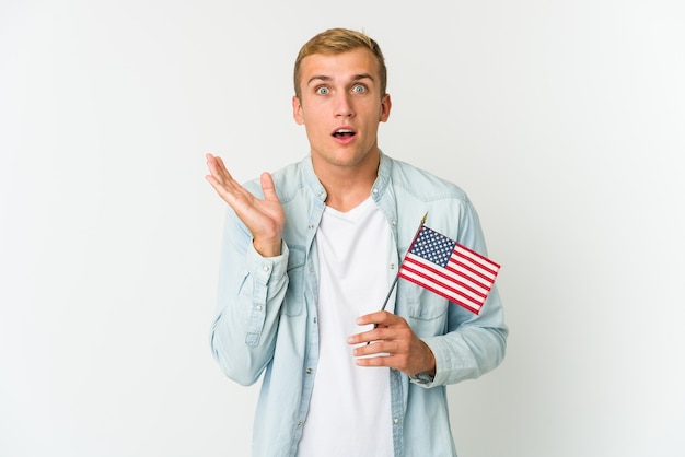 Hombre caucásico joven que sostiene una bandera americana aislada en blanco sorprendido y conmocionado.