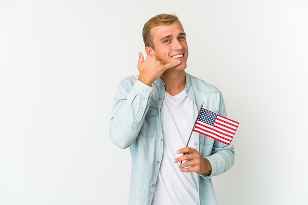 Hombre caucásico joven que sostiene una bandera americana aislada que muestra un gesto de llamada de teléfono móvil con los dedos.