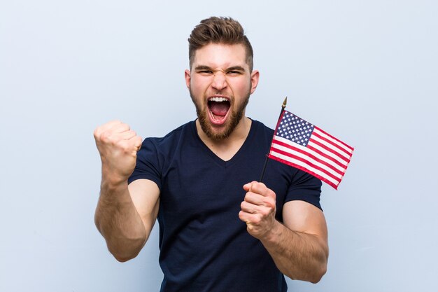 Hombre caucásico joven que sostiene una bandera de Estados Unidos animando despreocupado y emocionado. Concepto de victoria.