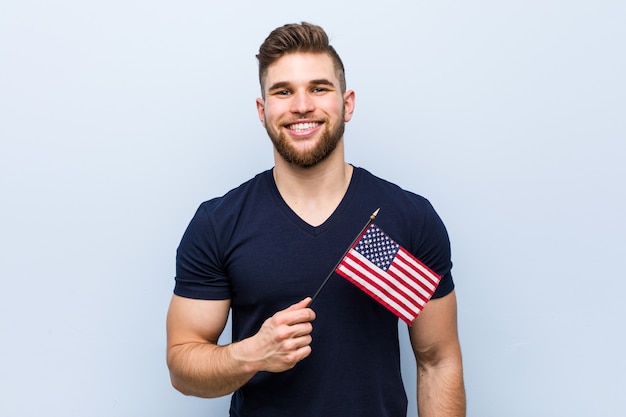 Hombre caucásico joven que sostiene una bandera de Estados Unidos feliz, sonriente y alegre.