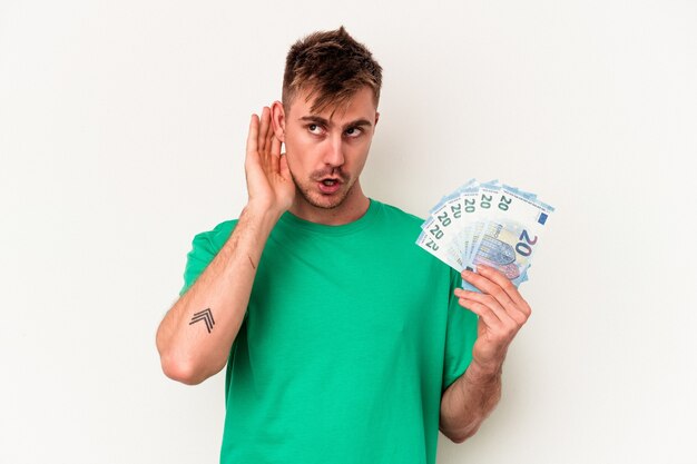 Hombre caucásico joven que sostiene los billetes de banco aislado en el fondo blanco que intenta escuchar un chisme.