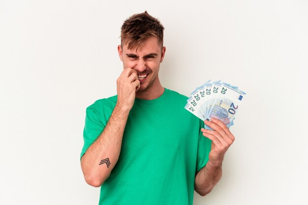 Hombre caucásico joven que sostiene los billetes de banco aislado en el fondo blanco que muerde las uñas, nervioso y muy ansioso.