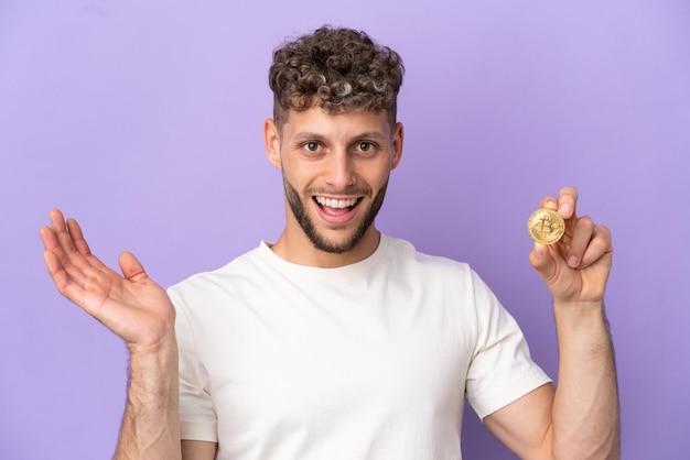 Hombre caucásico joven que sostiene un Bitcoin aislado sobre fondo púrpura con expresión facial sorprendida