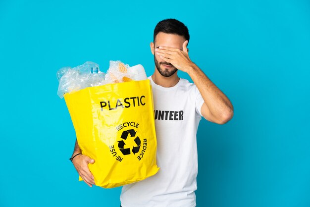 Hombre caucásico joven que sostiene una bolsa llena de botellas de plástico para reciclar aislado sobre fondo azul que cubre los ojos con las manos. No quiero ver algo