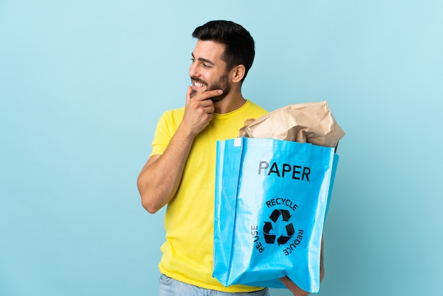 Hombre caucásico joven que sostiene una bolsa de reciclaje aislada en la pared azul que piensa una idea y que mira el lado