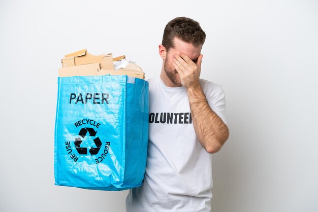 Hombre caucásico joven que sostiene una bolsa de reciclaje llena de papel para reciclar aislado sobre fondo blanco con expresión cansada y enferma