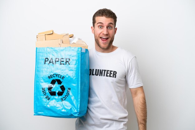 Hombre caucásico joven que sostiene una bolsa de reciclaje llena de papel para reciclar aislado sobre fondo blanco con expresión facial sorpresa