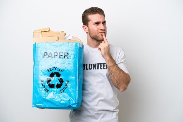 Hombre caucásico joven que sostiene una bolsa de reciclaje llena de papel para reciclar aislado sobre fondo blanco que tiene dudas mientras mira hacia arriba