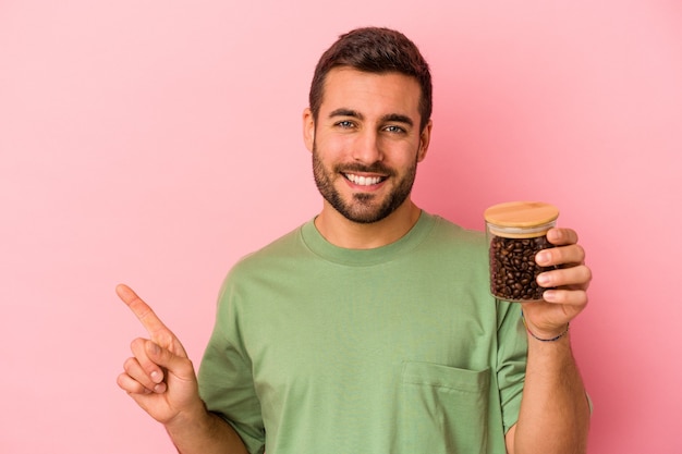 Hombre caucásico joven que sostiene una botella de café aislada en la pared rosada que sonríe y que señala a un lado, que muestra algo en el espacio en blanco