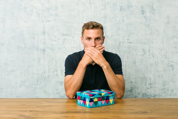 Hombre caucásico joven que sostiene una caja de regalo en una tabla sorprendida cubriendo la boca con las manos.