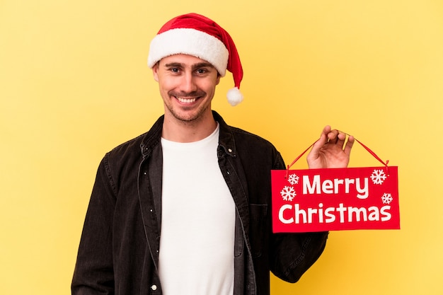 Hombre caucásico joven que sostiene un cartel de Feliz Navidad aislado sobre fondo amarillo feliz, sonriente y alegre.