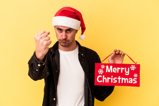 Hombre caucásico joven que sostiene un cartel de Feliz Navidad aislado sobre fondo amarillo que muestra el puño a la cámara, expresión facial agresiva.