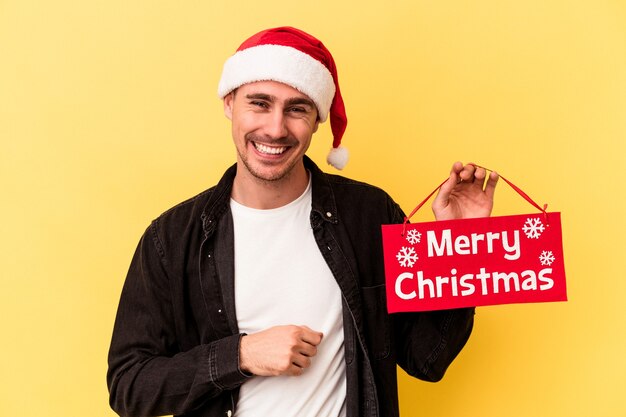 Hombre caucásico joven que sostiene un cartel de Feliz Navidad aislado sobre fondo amarillo riendo y divirtiéndose.