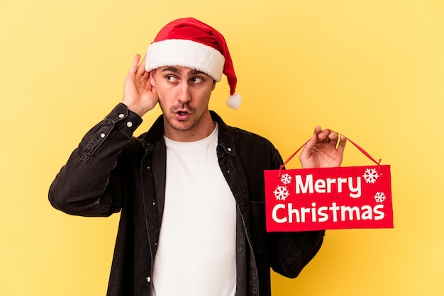 Hombre caucásico joven que sostiene un cartel de Feliz Navidad aislado sobre fondo amarillo tratando de escuchar un chisme.