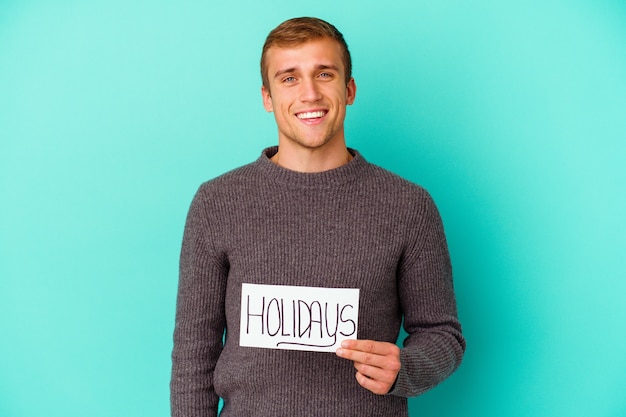 Hombre caucásico joven que sostiene un cartel de vacaciones aislado sobre fondo azul feliz, sonriente y alegre.