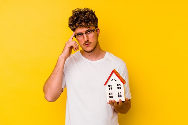 Hombre caucásico joven que sostiene una casa modelo aislada sobre fondo amarillo señalando el templo con el dedo, pensando, se centró en una tarea.