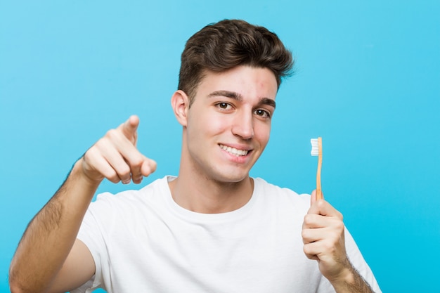 Hombre caucásico joven que sostiene un cepillo de dientes joven mujer india vistiendo un pijama y máscara para dormir aislado sosteniendo una almohada sonrisas alegres apuntando al frente. <mixto>