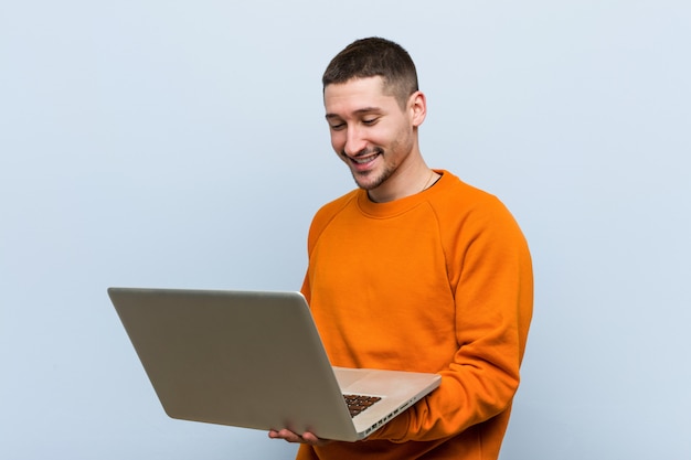 Hombre caucásico joven que sostiene una computadora portátil feliz, sonriente y alegre.