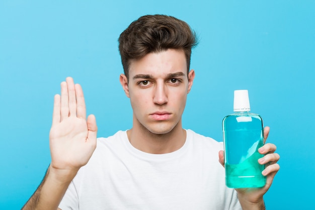 Hombre caucásico joven que sostiene un enjuague bucal de pie con la mano extendida mostrando la señal de stop, impidiéndote.