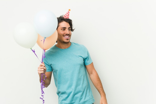 Hombre caucásico joven que sostiene los globos que celebran un cumpleaños aislado en una pared gris