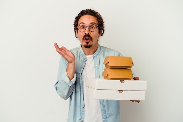 Hombre caucásico joven que sostiene una hamburguesa pizzas aisladas sobre fondo blanco sorprendido y conmocionado.