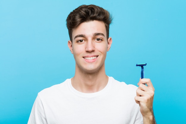 Hombre caucásico joven que sostiene una hoja de afeitar feliz, sonriente y alegre.