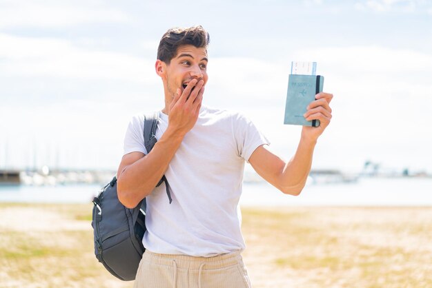 Hombre caucásico joven que sostiene un pasaporte al aire libre con sorpresa y expresión facial conmocionada