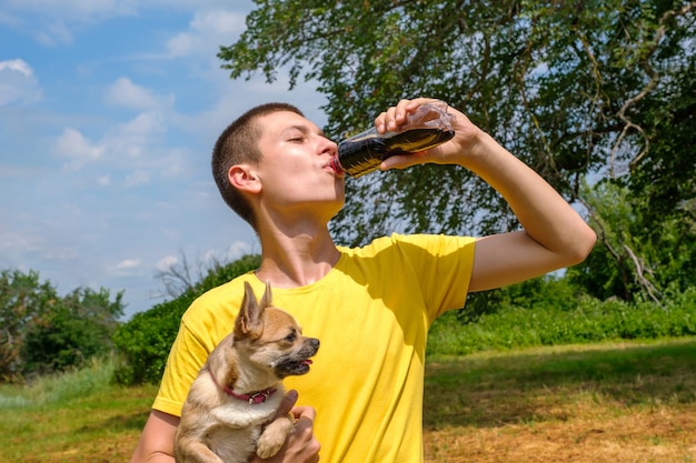 Hombre caucásico joven que sostiene el perro de la chihuahua y que bebe cola de la botella al aire libre. Caminando en el parque en verano, vista de ángulo bajo