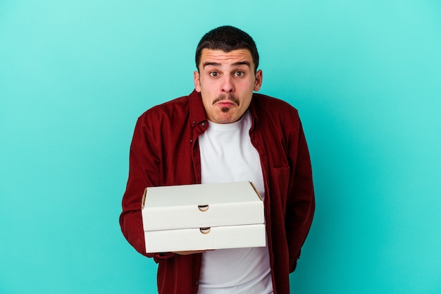 Hombre caucásico joven que sostiene pizzas aisladas en la pared azul se encoge de hombros y abre los ojos confundidos.