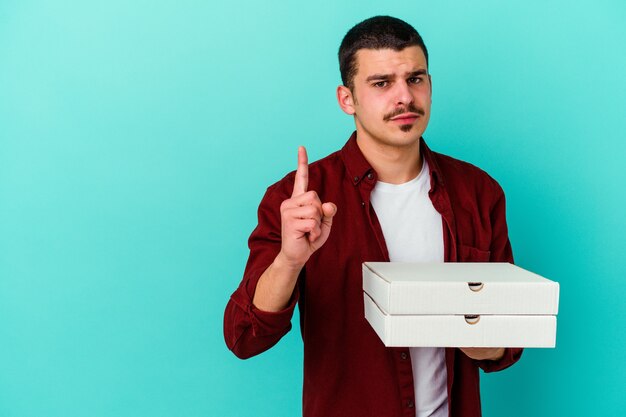 Hombre caucásico joven que sostiene las pizzas aisladas sobre fondo azul que muestra el número uno con el dedo.