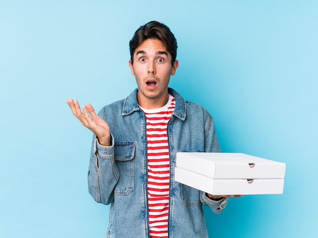 Hombre caucásico joven que sostiene las pizzas aisladas sorprendido y conmocionado.