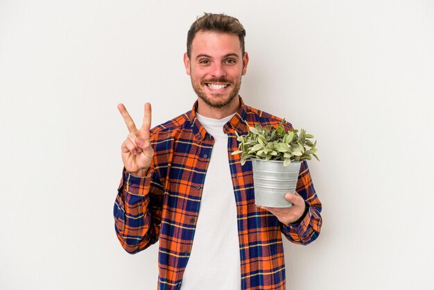 Hombre caucásico joven que sostiene una planta aislada en el fondo blanco que muestra el número dos con los dedos.