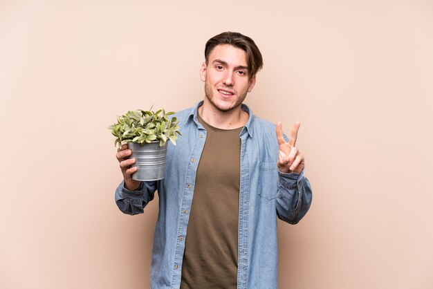 Hombre caucásico joven que sostiene una planta que muestra el número dos con los dedos.