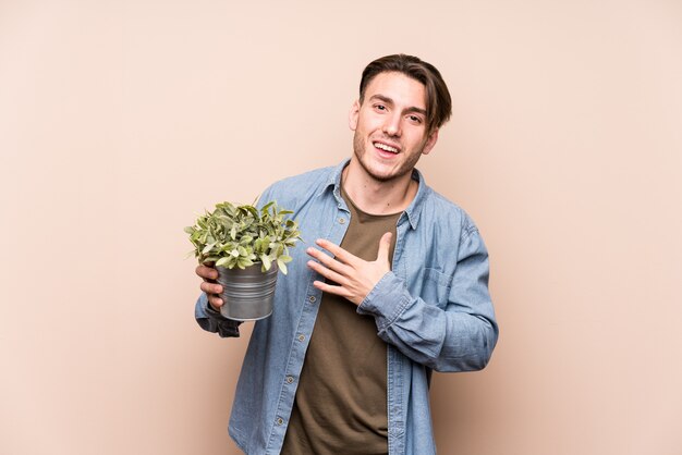 El hombre caucásico joven que sostiene una planta ríe a carcajadas manteniendo la mano en el pecho.