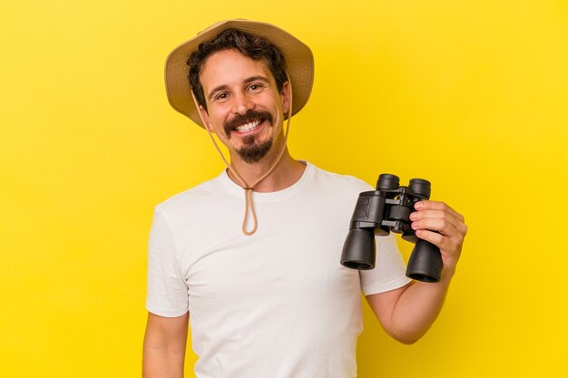 Hombre caucásico joven que sostiene los prismáticos aislados sobre fondo amarillo feliz, sonriente y alegre.