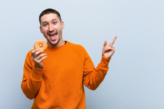 Hombre caucásico joven que sostiene una rosquilla que sonríe alegremente que señala con el índice lejos.