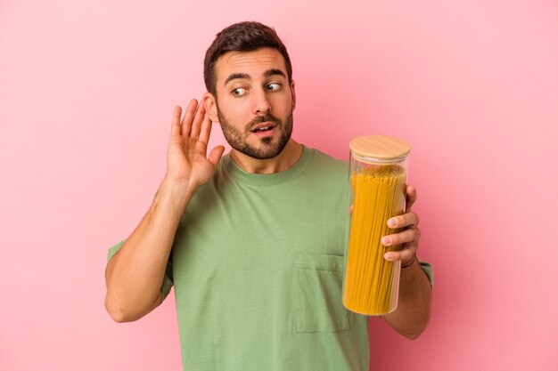 Hombre caucásico joven que sostiene el tarro de pasta aislado en la pared rosada tratando de escuchar un chisme.