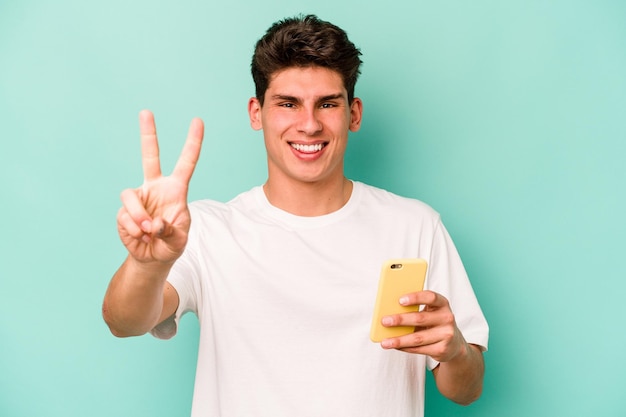 Hombre caucásico joven que sostiene el teléfono móvil aislado en el fondo azul que muestra el número dos con los dedos