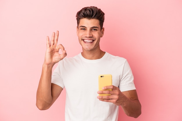 Hombre caucásico joven que sostiene un teléfono móvil aislado en fondo rosa alegre y confiado que muestra el gesto aceptable.