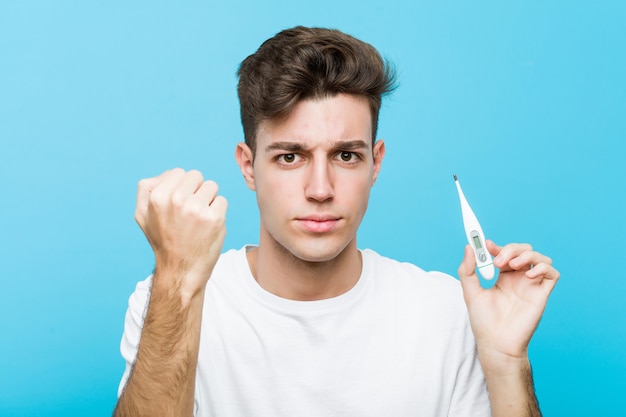 Hombre caucásico joven que sostiene un termómetro médico que muestra el puño a la cámara, expresión facial agresiva.