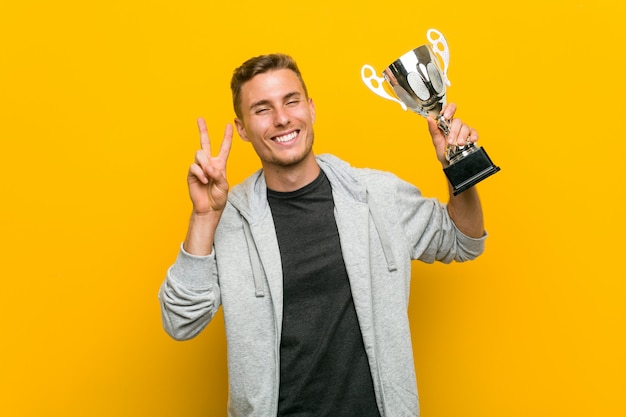 Foto hombre caucásico joven que sostiene un trofeo que muestra la muestra de la victoria y que sonríe ampliamente.