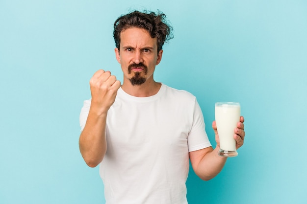 Hombre caucásico joven que sostiene un vaso de leche aislado sobre fondo azul que muestra el puño a la cámara, expresión facial agresiva.