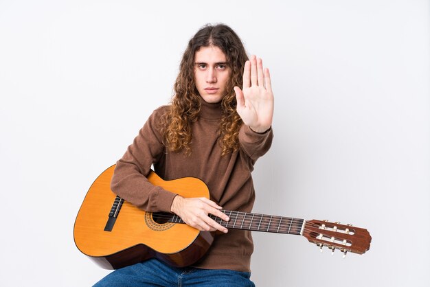 Hombre caucásico joven que toca la guitarra aislada que se coloca con la mano extendida que muestra la señal de pare, previniéndole.