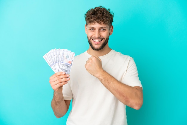 Hombre caucásico joven que toma un montón de dinero aislado sobre fondo azul celebrando una victoria