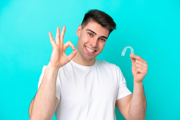 Hombre caucásico joven que usa aparatos invisibles aislados en fondo azul que muestra el signo de ok con los dedos