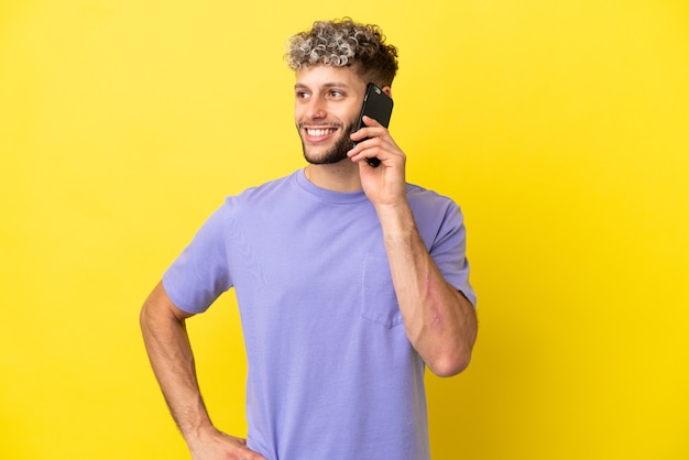 Hombre caucásico joven que usa el teléfono móvil aislado en fondo amarillo que presenta con los brazos en la cadera y que sonríe
