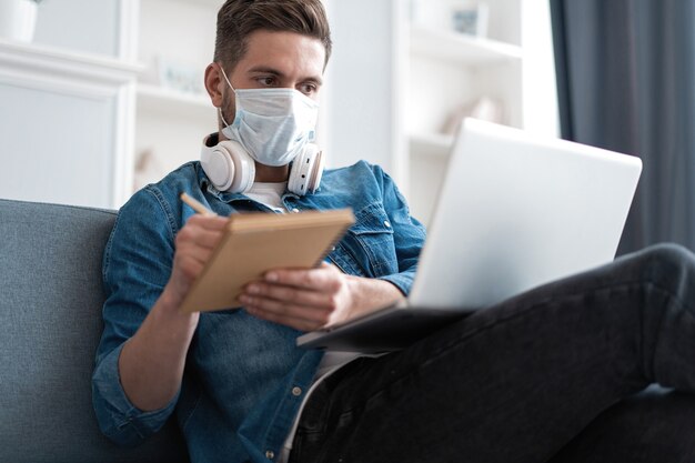 Hombre caucásico joven quedarse en casa, trabajar desde casa, usar máscara protectora, usar la computadora portátil e internet. Pandemia de coronavirus, cuarentena del covid 19