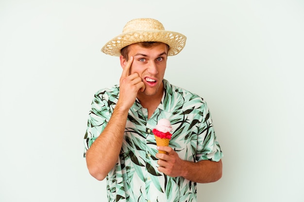 Hombre caucásico joven con ropa de verano y sosteniendo un helado aislado en blanco mostrando un gesto de decepción con el dedo índice.