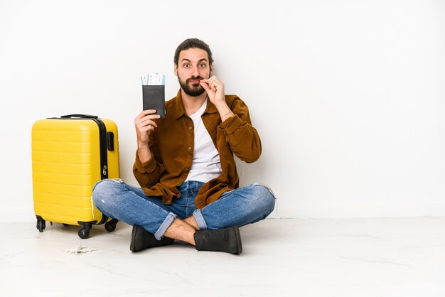 Hombre caucásico joven sentado sosteniendo un pasaporte y una maleta aislada con los dedos en los labios manteniendo un secreto.