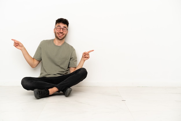 Hombre caucásico joven sentado en el suelo aislado sobre fondo blanco que señala con el dedo a los laterales y feliz